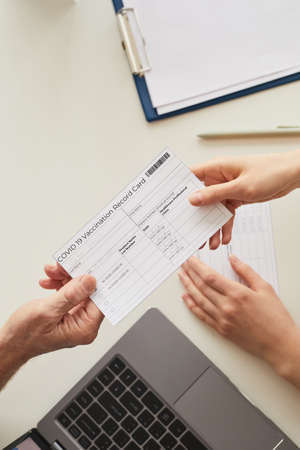 Top view close up of doctor giving Covid vaccination card to patient across table in clinic, copy...