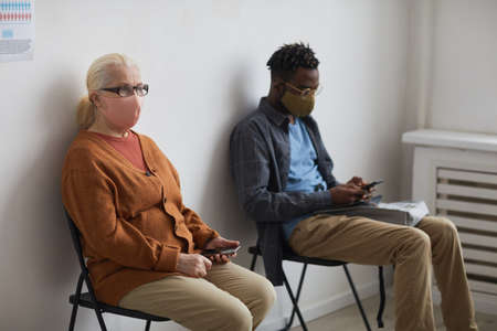 Portrait of two people wearing masks and sitting on chairs while waiting in line at clinic, copy...