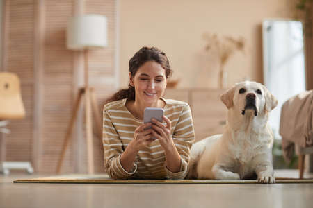 Front view portrait of smiling young woman lying on floor with dog and using smartphone in cozy home...