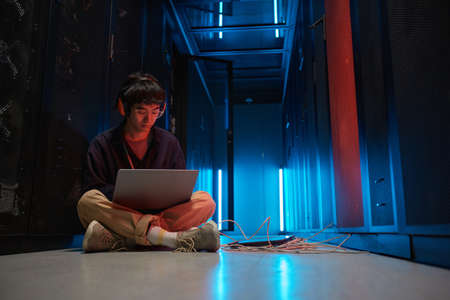Full length portrait of young Asian man using laptop while sitting on floor in server room lit by...