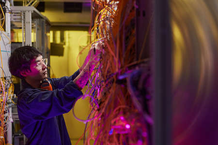 Wide angle portrait of young network technician connecting wires and cables in server room, copy...