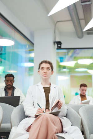 Vertical front view portrait of young woman wearing lab coat while sitting in audience and listening to lecture on medicine in college or coworking centerの写真素材
