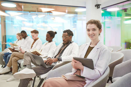 Portrait of young woman wearing lab coat and smiling at camera while sitting in row with multi-ethnic group of people in audience at medical seminar, copy spaceの写真素材