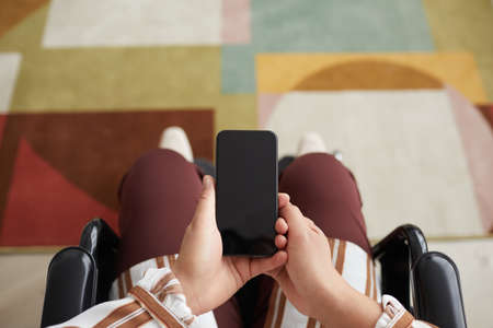 Top view close up of unrecognizable disabled woman holding smartphone while sitting in wheelchair, copy spaceの写真素材