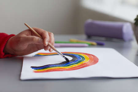 Close up of unrecognizable woman painting rainbow as LGBTQ symbol while sitting at desk at home, focus on hand holding paintbrush, copy spaceの写真素材