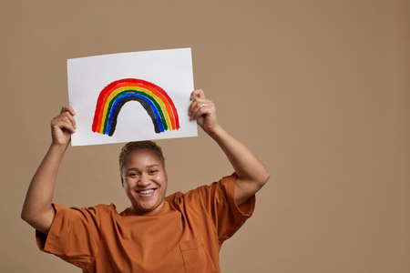 Portrait of carefree mixed-race woman holding rainbow sign and smiling at camera while posing in studioの写真素材