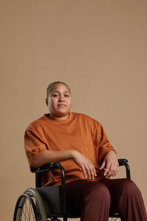 Vertical portrait of contemporary mixed race woman using wheelchair and looking at camera while posing in studio against neutral backgroundの写真素材