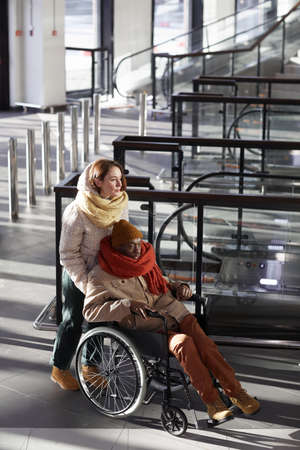 Vertical full length portrait of young woman assisting African American man in wheelchair at subway...