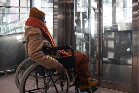 Full length portrait of African American man in wheelchair using accessible elevator in urban city...