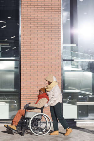 Vertical full length portrait of African American man in wheelchair moving in urban city with young...