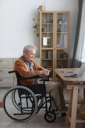 Vertical side view portrait of senior man in wheelchair using smartphone while sitting by desk in home interiorの写真素材