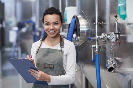 Waist up portrait of young African-American woman enjoying work at industrial factory and smiling at...