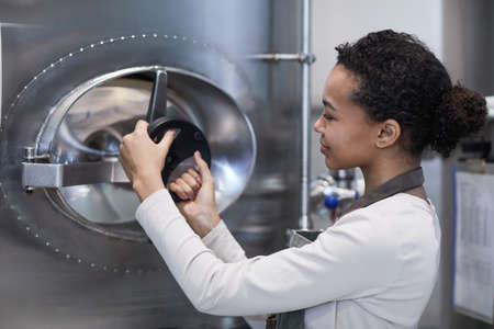 Side view portrait of smiling female worker opening hatch at storage tank in industrial workshop, copy spaceの写真素材