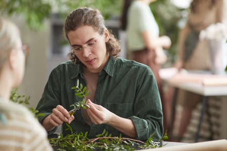 Portrait of young male florist cutting green plants while creating floral compositions for decor, copy spaceの写真素材
