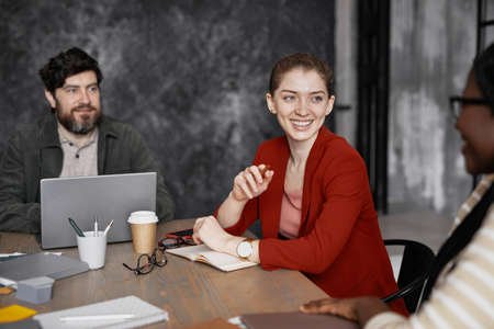 Portrait of smiling young businesswoman wearing red jacket during meeting with coworkers in office, copy spaceの写真素材