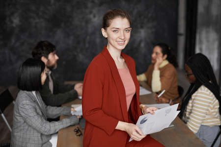 Waist up portrait of successful young businesswoman wearing red jacket and smiling at camera with diverse group of people meeting in background, copy spaceの写真素材