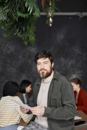 Vertical waist up portrait of contemporary bearded businessman looking at camera with diverse group of people meeting in backgroundの写真素材