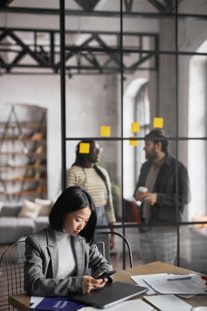 Vertical portrait of elegant Asian businesswoman working at table in graphic grey office interior,...