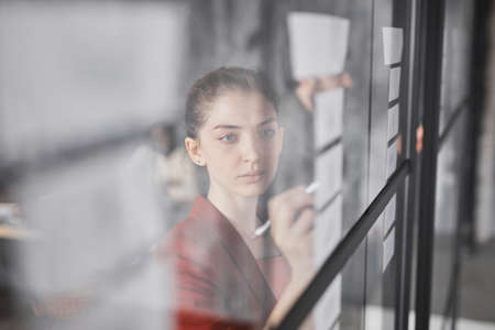 Portrait of young businesswoman writing on glass wall while planning project in modern office, copy spaceの写真素材