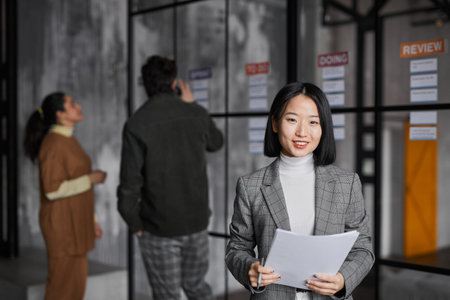 Waist up portrait of smiling Asian businesswoman standing by glass wall while planning project in modern office, copy spaceの写真素材