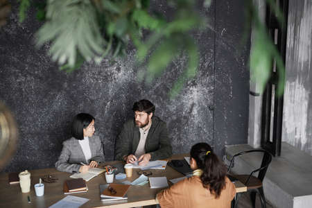 Portrait of bearded businessman talking to female colleagues during meeting at table in minimal grey office, copy spaceの写真素材