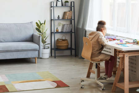 Full length portrait of cute African-American girl doing homework or drawing while sitting at desk in cozy home interior, copy spaceの写真素材