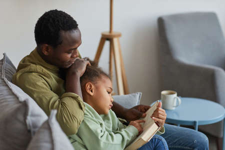 Side view portrait of loving African-American father and daughter reading while sitting on couch...