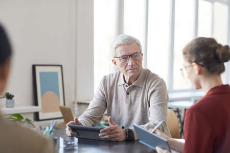 Portrait of white haired senior businessman listening to female colleague during briefing meeting in officeの写真素材