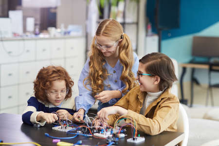 Portrait of three smiling children experimenting with electric circuits while building robots in...