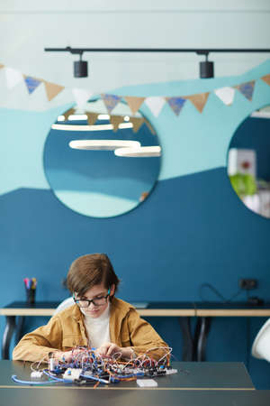 Vertical portrait of cute boy working with electric wires while building robot in engineering class,...