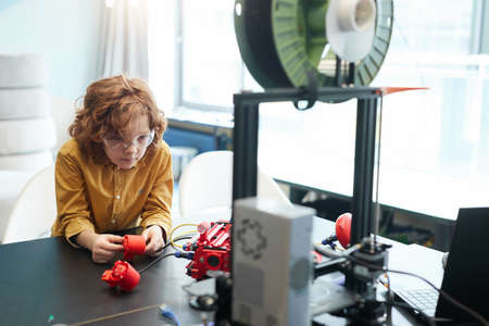 Portrait of cute curly haired boy building robot boat during engineering class at modern school,...