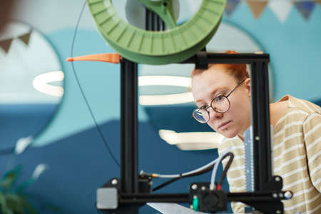 Framed portrait of young red haired woman using 3D printer in engineering class, copy space
