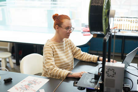 High angle portrait of young red haired woman using 3D printer during engineering class at college,...