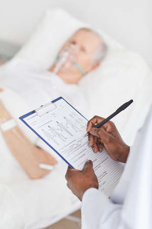 Close up of African-American doctor holding clipboard while standing by patients bed in hospital, copy spaceの写真素材
