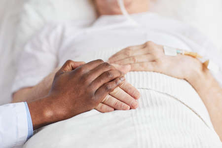 Close up of caring African-American doctor holding hands with senior patient lying in hospital bed,...