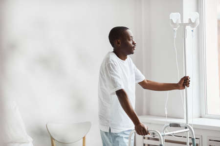 Side view portrait of African-American man in hospital holding onto iv drip stand and looking at...