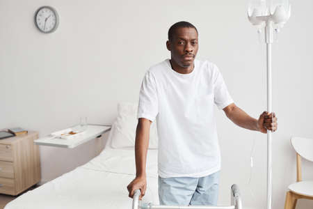 Waist up portrait of African-American man in hospital holding onto iv drip stand and looking at camera, copy spaceの写真素材