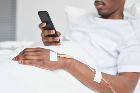 Close up of African-American man in hospital bed and using smartphone with focus on iv drip catheter in hand, copy spaceの写真素材