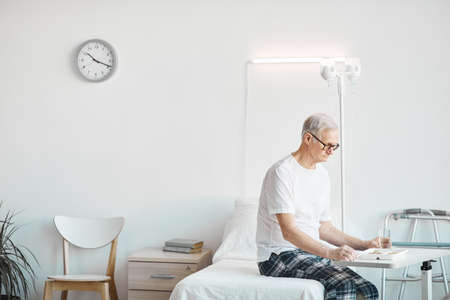Side view portrait of senior man eating food in hospital while sitting on bed in white room, copy...