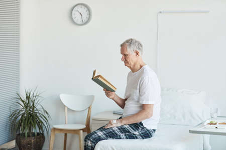Side view portrait of senior man reading book in hospital while sitting on bed in white room copy...