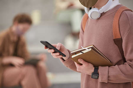 Cropped portrait of unrecognizable male student holding smartphone and wearing mask in school interior, copy spaceの写真素材