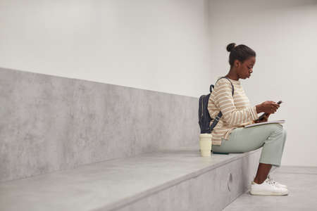 Graphic side view portrait of female African-American student using smartphone while relaxing in...