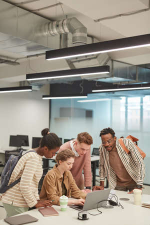Vertical wide angle shot of diverse group of young students using laptop together while working on...
