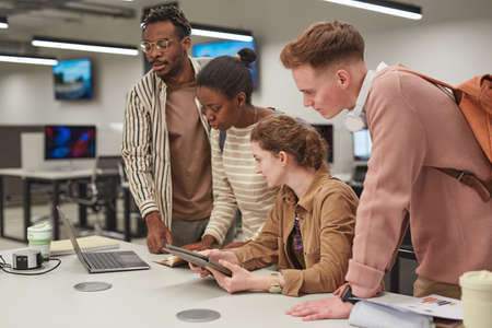 Diverse group of students working together and using computers while standing at table in school IT...