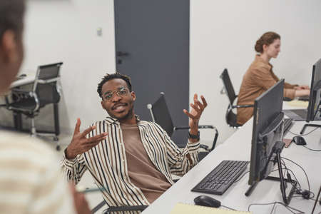 Portrait of African-American man talking to colleague in office and gesturing actively while sitting...