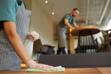 Close up of unrecognizable female waitress cleaning table in coffee shop with warm wooden accents,...