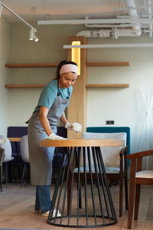 Vertical full length portrait of young mixed-race waitress cleaning table in cafe or coffee shop...