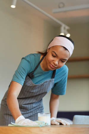 Vertical portrait of young mixed-race waitress cleaning table in cafe or coffee shop while preparing...