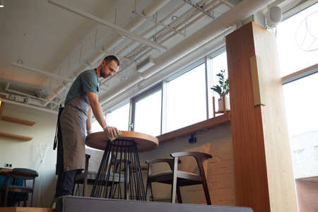 Low angle portrait of male waiter cleaning tables in cafe or coffee shop while preparing for opening...