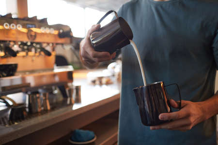 Cropped portrait of young barista pouring cream in latte while making fresh coffee in cafe or coffee...